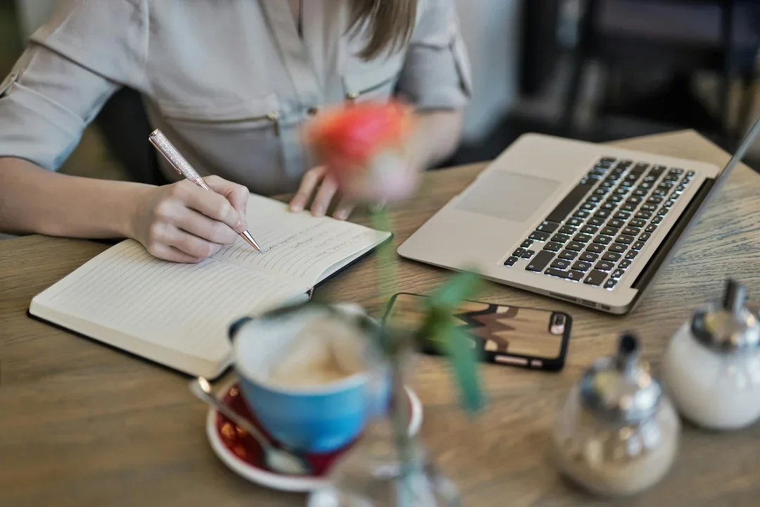 Person taking notes beside a laptop and a cup of coffee